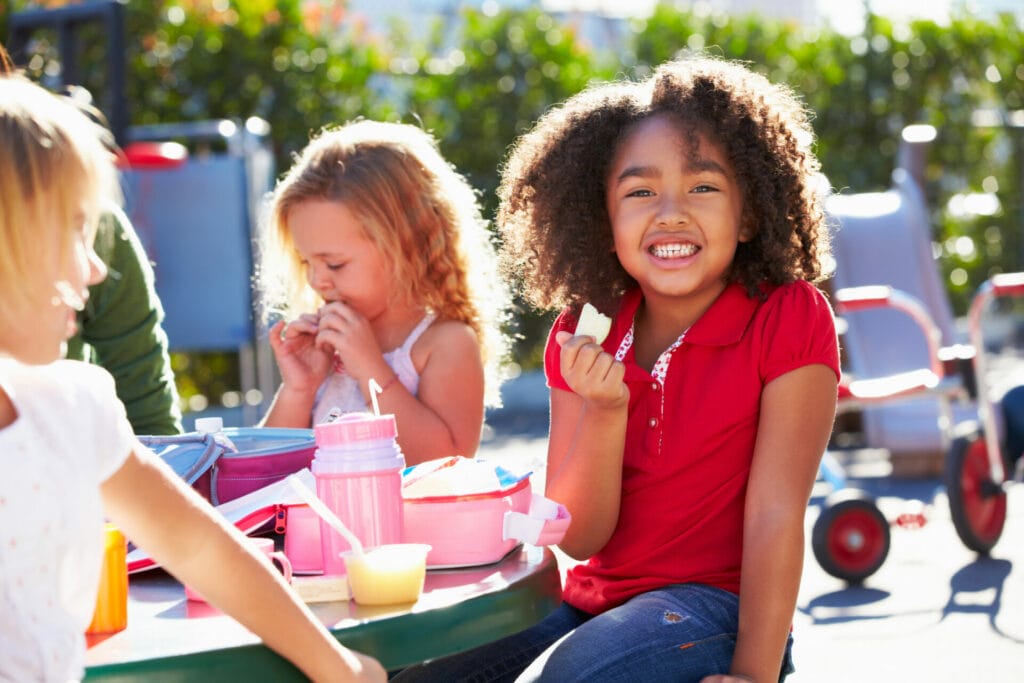 Elementary Children Sitting At Table Eating Lunch and Smiling