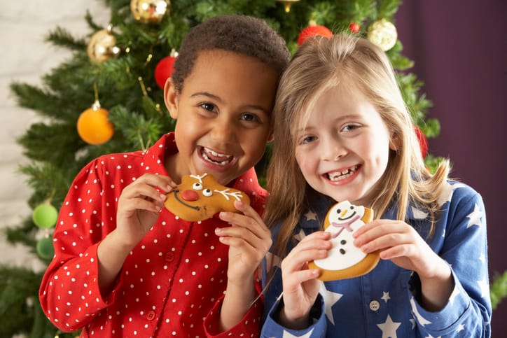 Two Young Children Eating Christmas Treats In Front Of Christmas Tree