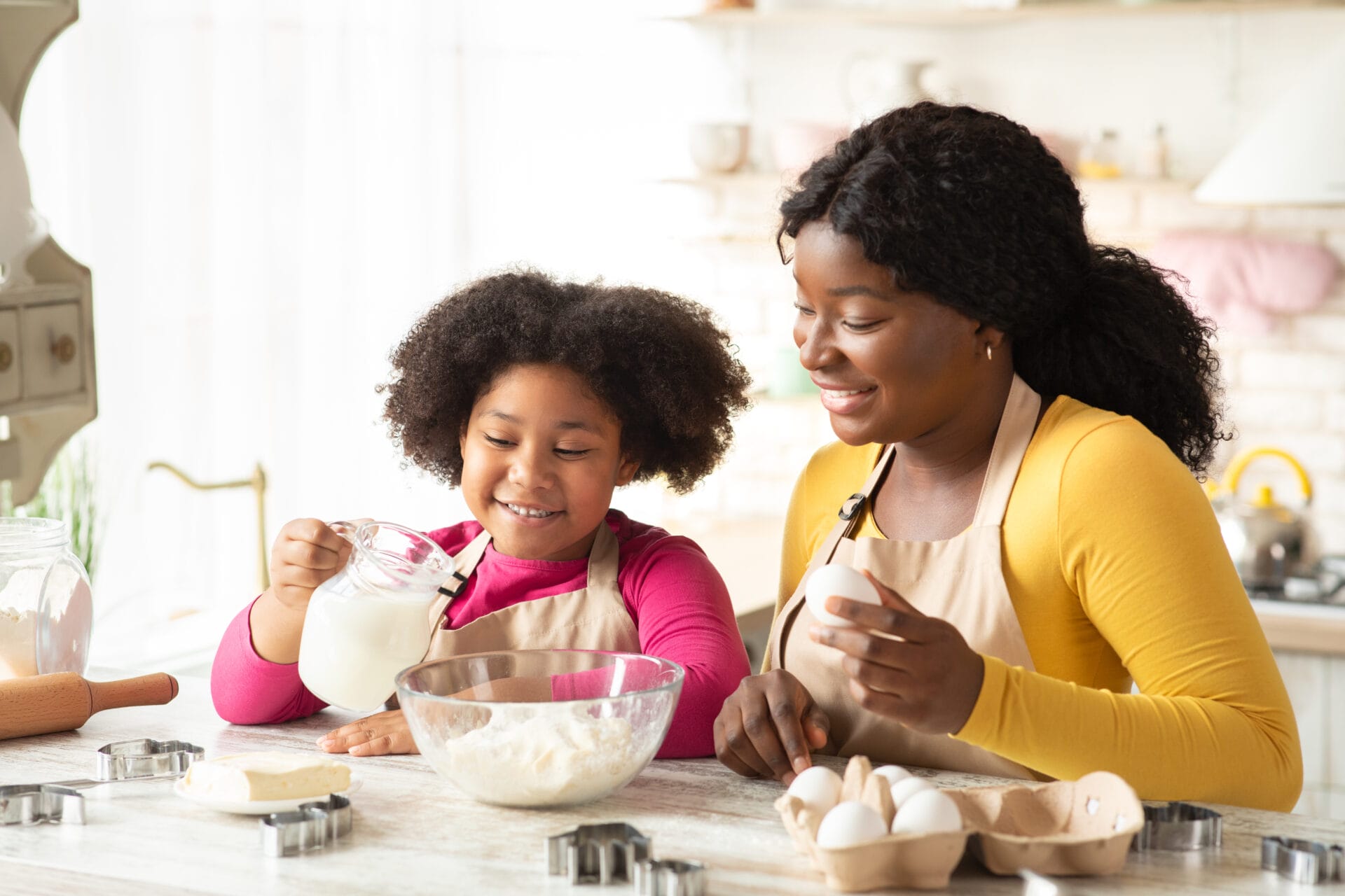 Mother and young daughter baking together in a bright kitchen, with the child pouring milk into a bowl of flour while the mother holds an egg and smiles supportively.