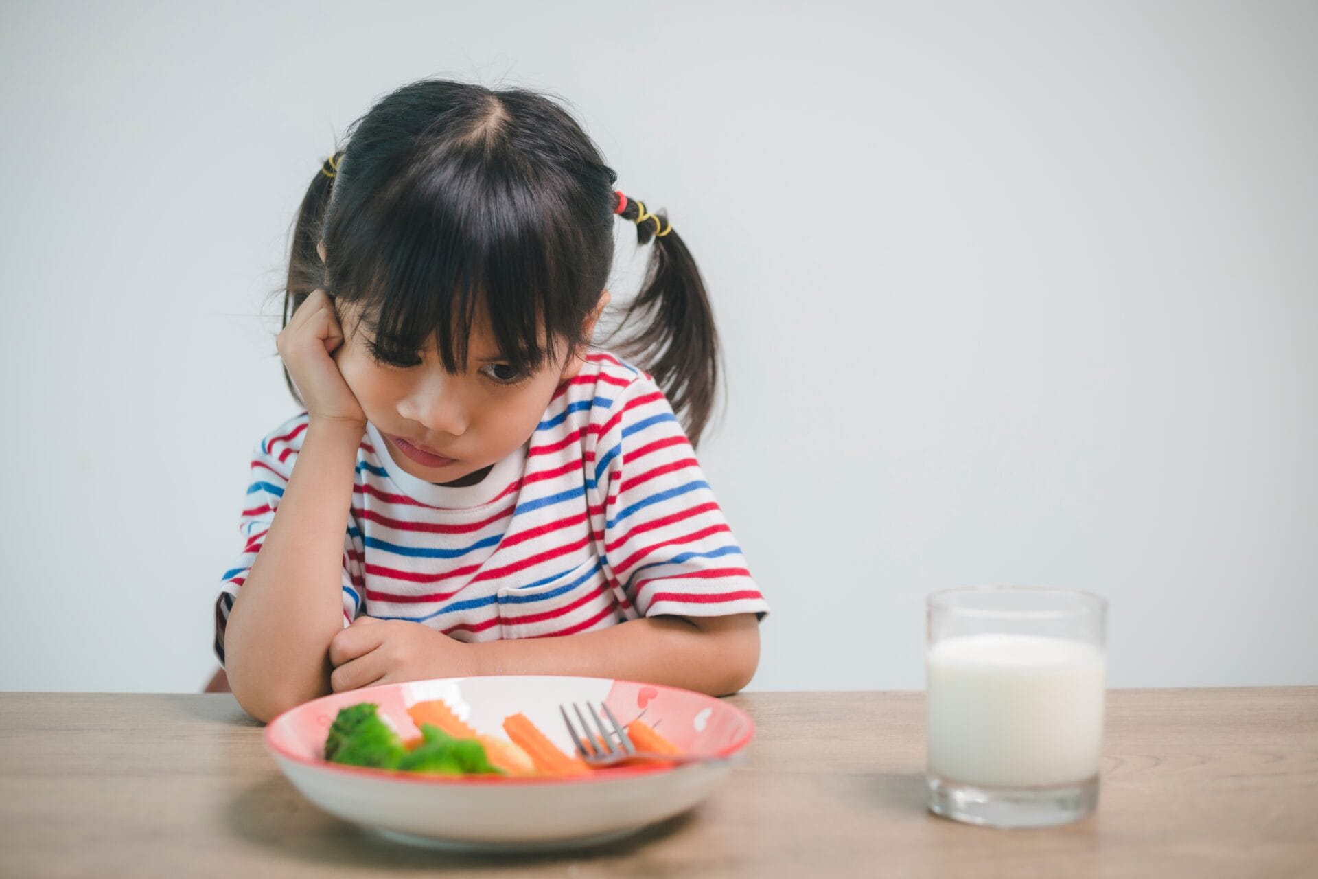 A young girl sits at the kitchen table begrudgingly with her hand on her face, a colorful plate of vegetables sits untouched in front of her.