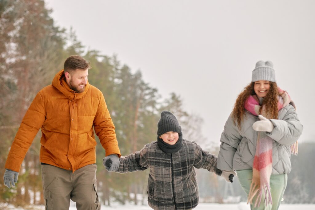 Man, woman and child walking together outdoors in winter, holding hands and smiling, snow covered trees in background.