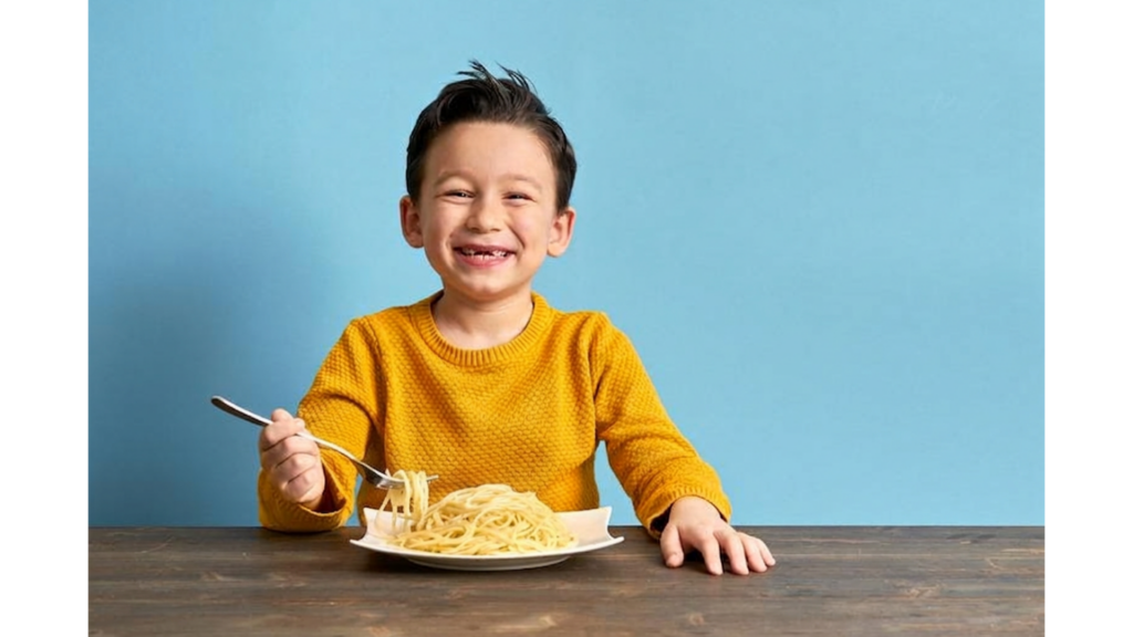 A young child smiling while eating a bowl of plain white pasta, illustrating the common "white foods only" phase in picky eaters.
